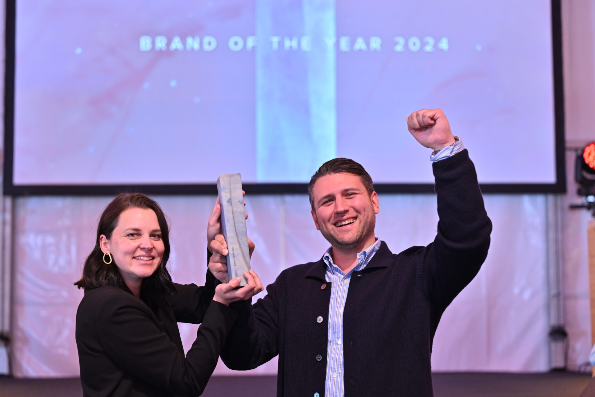 Eine Frau und ein Mann halten den Award "Brand of the Year 2024" in der Hand. Sie stehen vor einem hellen verschwommenen Hintergrund